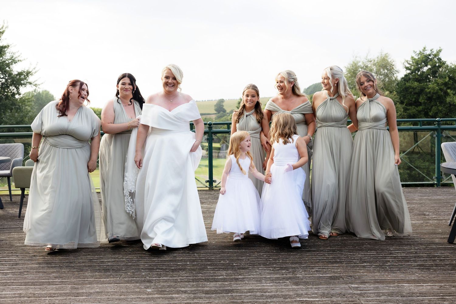 Bride in white gown poses with bridesmaids in sage green dresses and flower girls in white on a wooden deck.