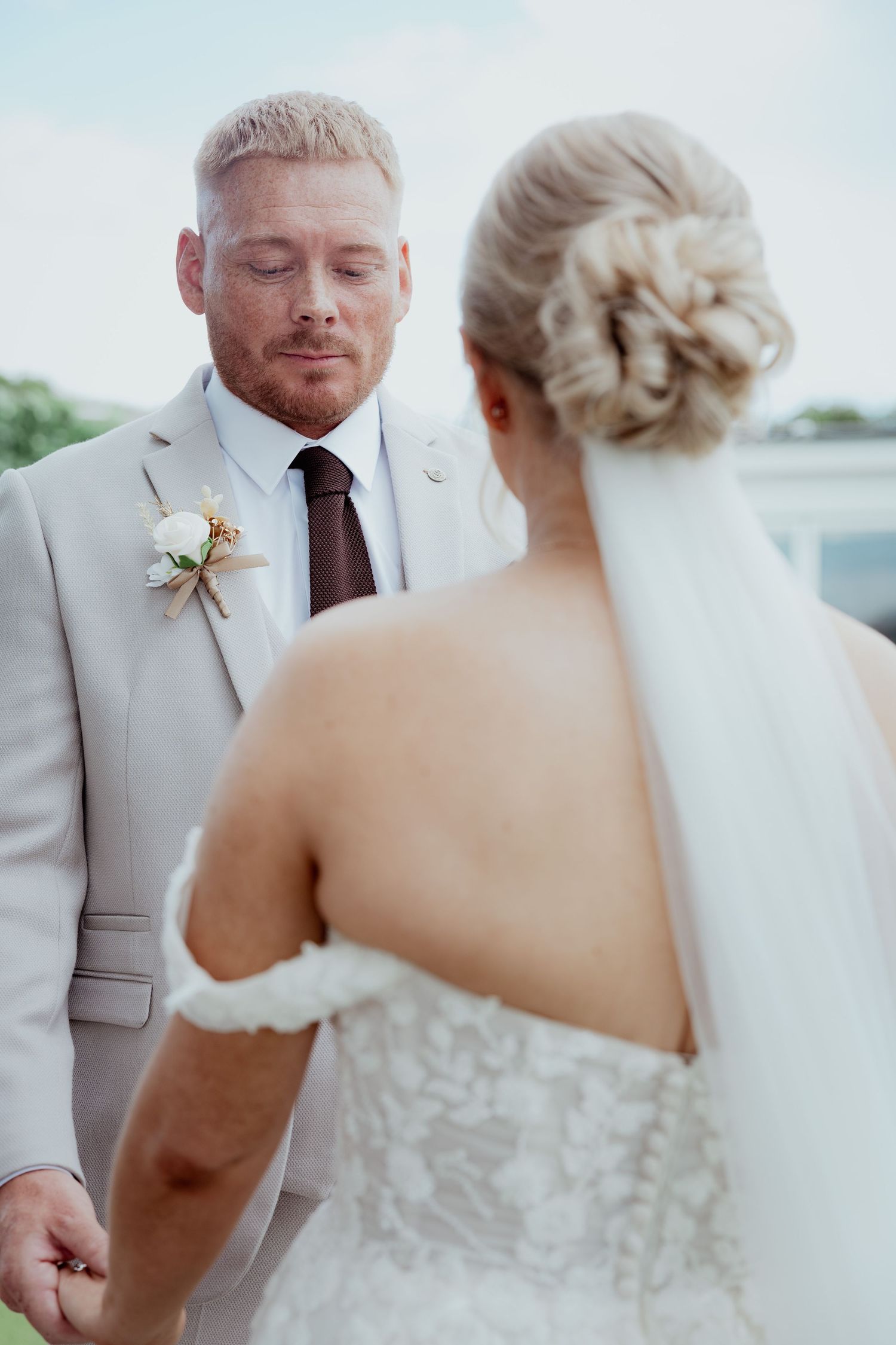 Emotional groom in beige suit with boutonniere holding brides hands during outdoor wedding ceremony.