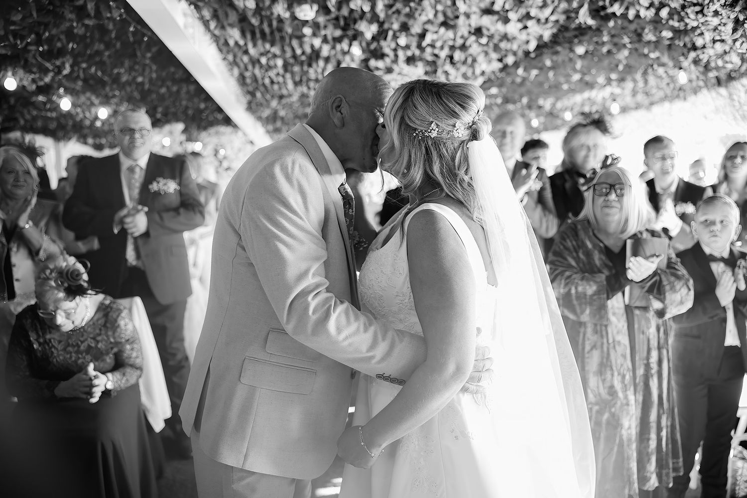 Black and white photo of bride and groom sharing first kiss surrounded by wedding guests.