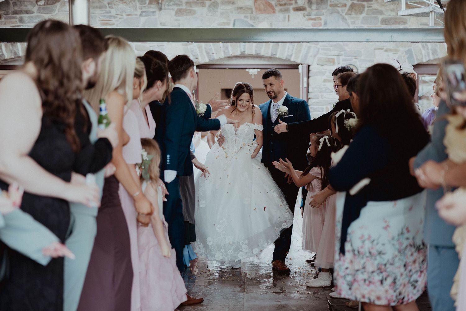 Bride and groom walk through confetti tunnel created by cheering guests at wedding reception.