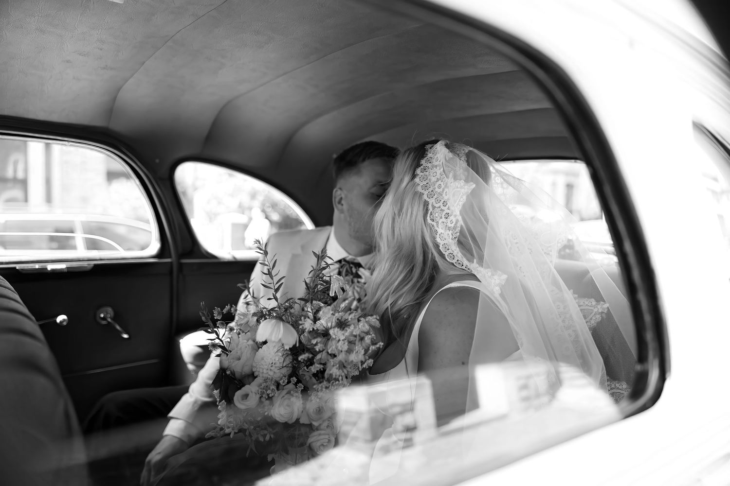 Romantic black and white photo of newlyweds sharing a kiss inside a vintage wedding car.