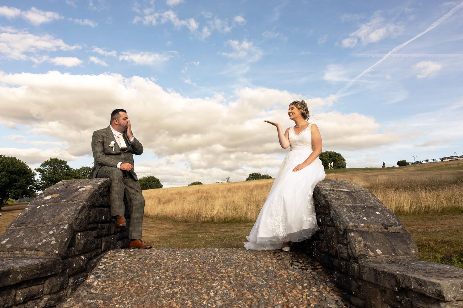 Bride and groom share a playful moment seated on stone walls in a scenic countryside wedding photo.