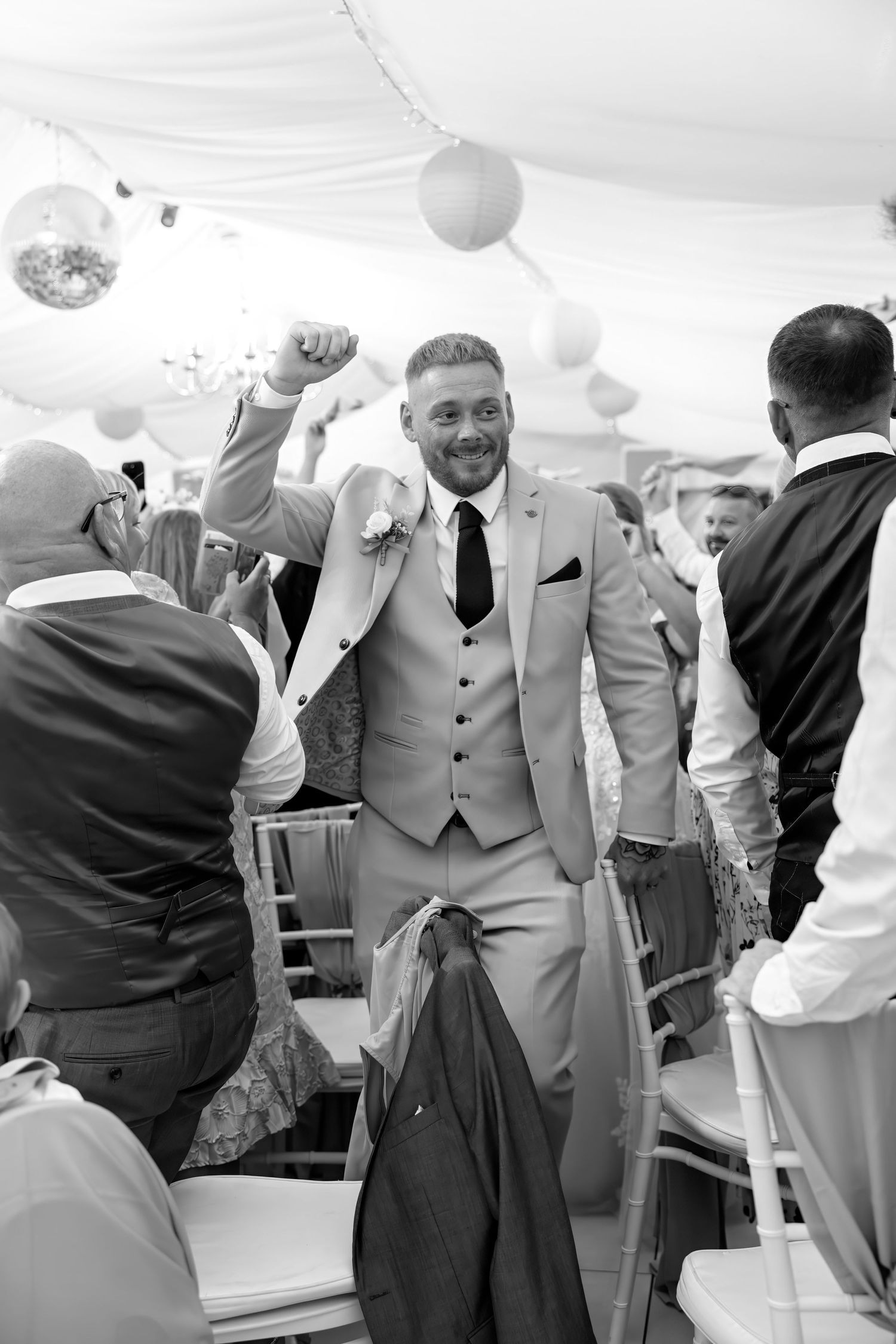 Smiling groom in light gray suit raises fist triumphantly while walking down aisle at elegant wedding reception.