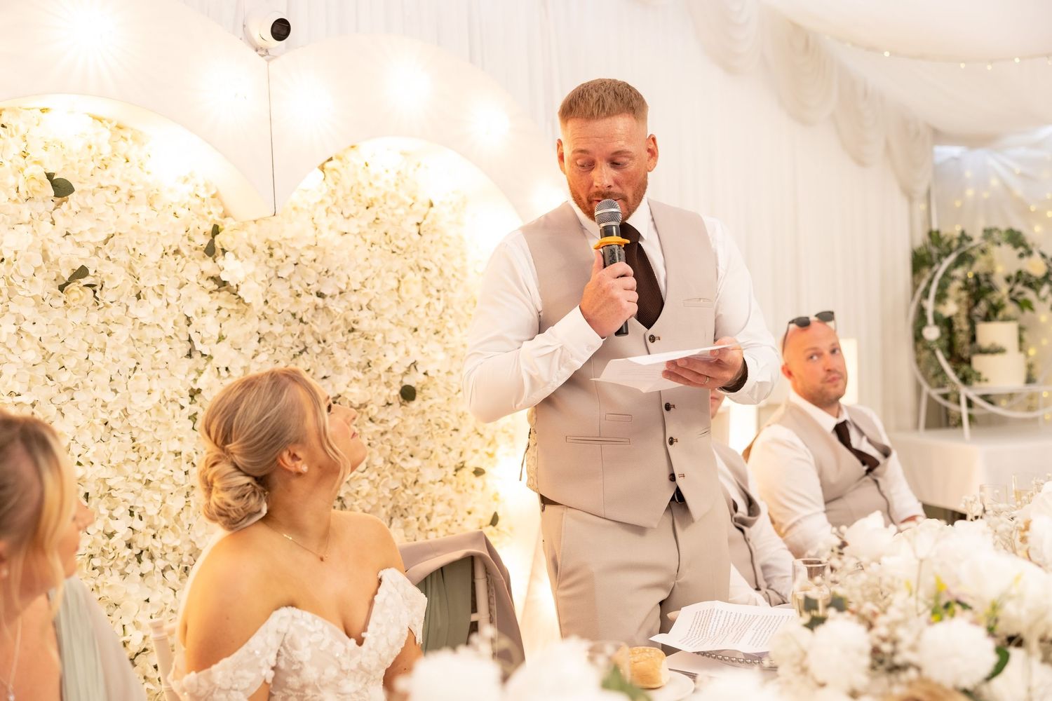 Best man reads wedding speech at beautifully decorated white floral reception venue.