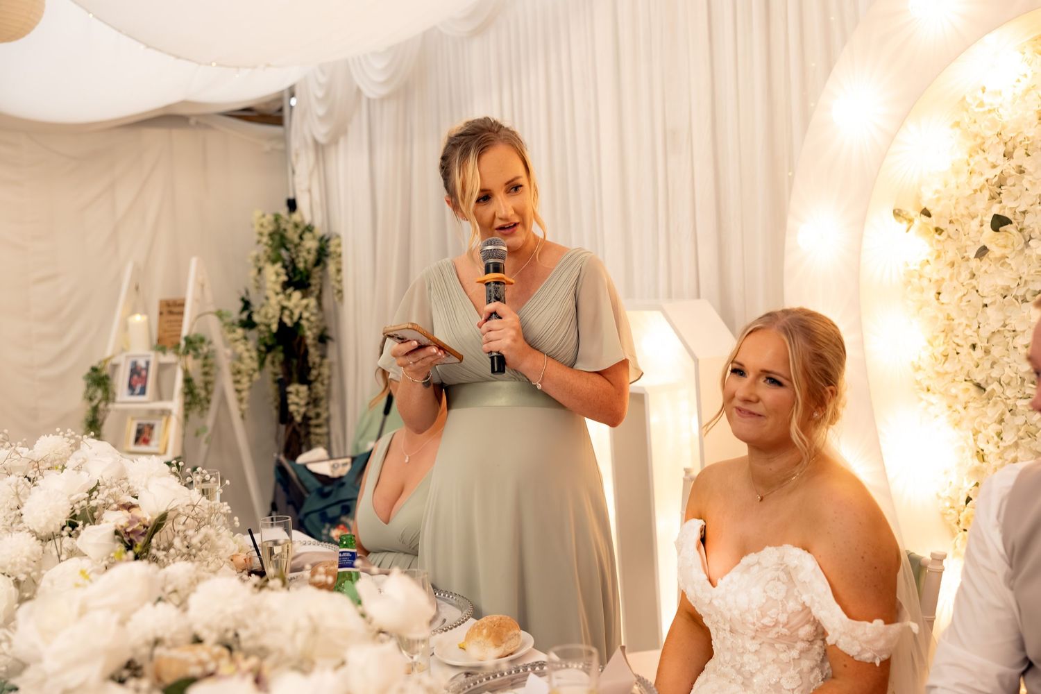 Bridesmaid in sage green dress gives a heartfelt speech at an elegant white floral wedding reception.
