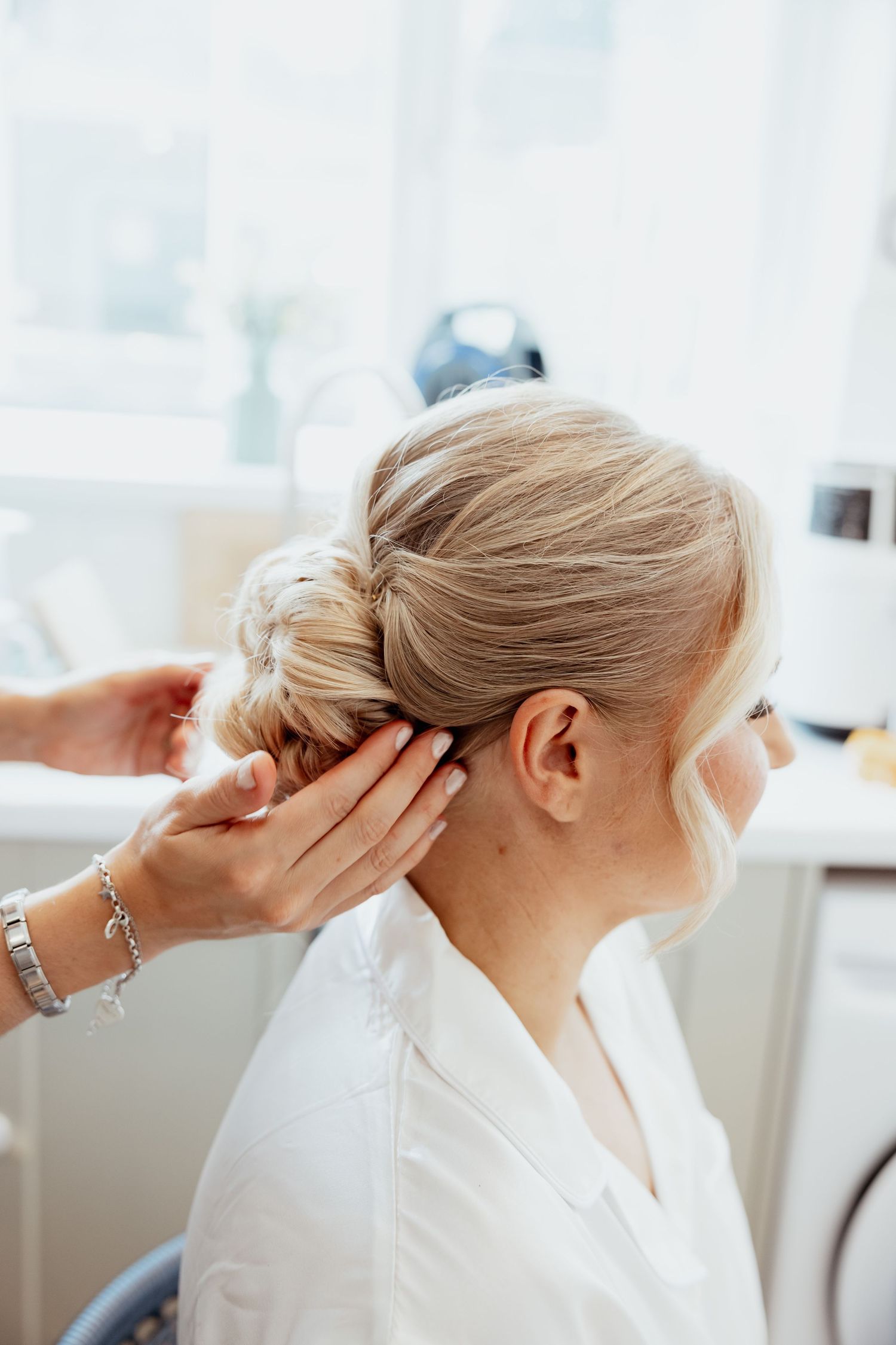 Hairstylist finishing an elegant bridal updo on a blonde bride wearing a white robe.