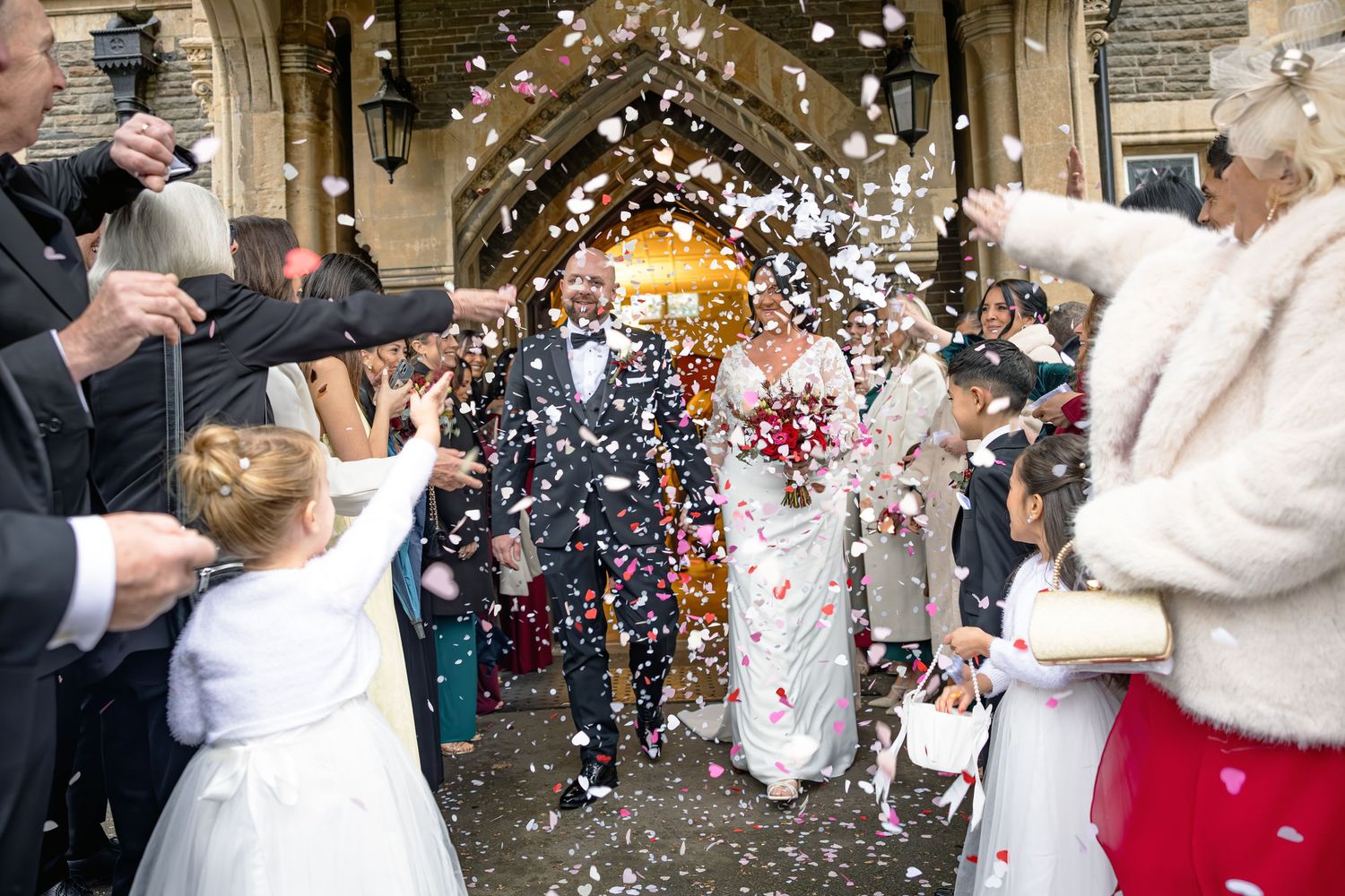 Bride and groom exit church through confetti shower, flanked by guests and flower girls in white dresses.