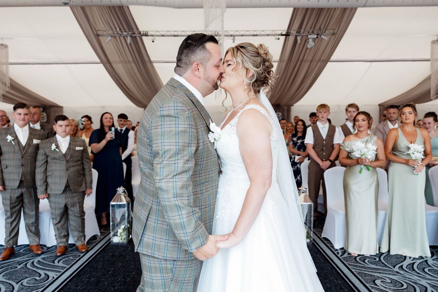 Bride and groom share first kiss at wedding ceremony with bridesmaids and guests applauding in elegant venue.