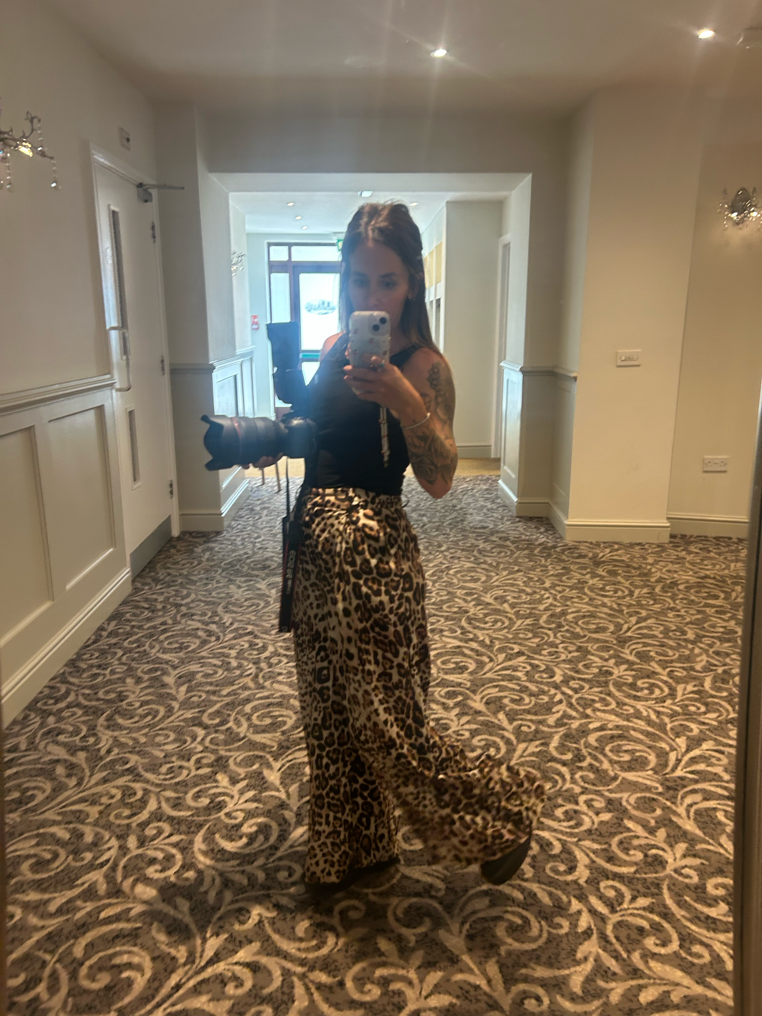 Woman in black top and leopard print maxi skirt taking mirror selfie in elegant hotel hallway.
