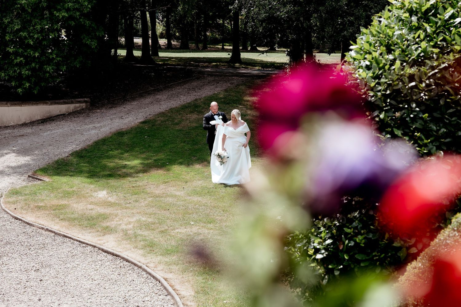 Bride and groom walking hand in hand through lush garden path with colorful roses in foreground.