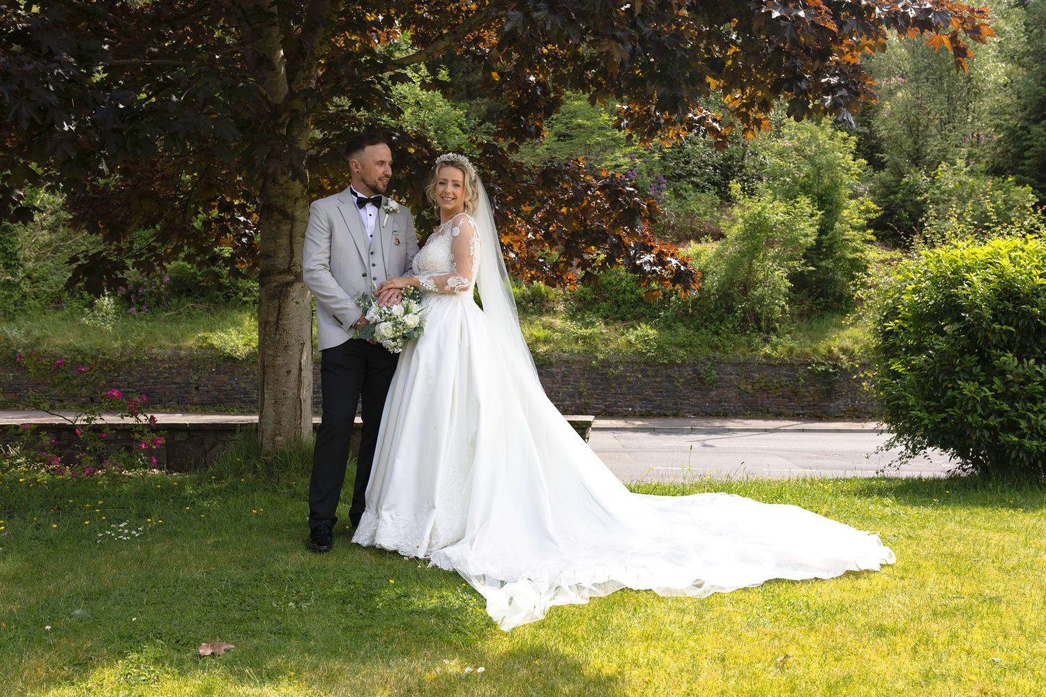Bride and groom pose together in lush  garden, bride wearing elegant white gown with long train, groom in grey suit. Heritage park hotel.
