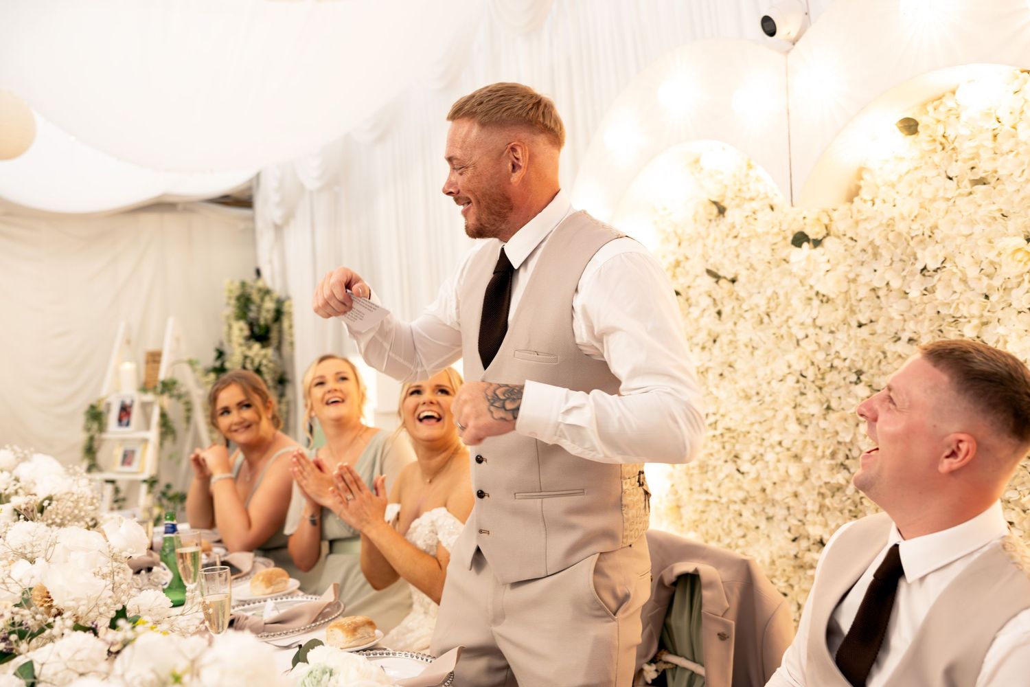 Energetic groomsman celebrates during wedding speeches as bride and guests laugh at reception.