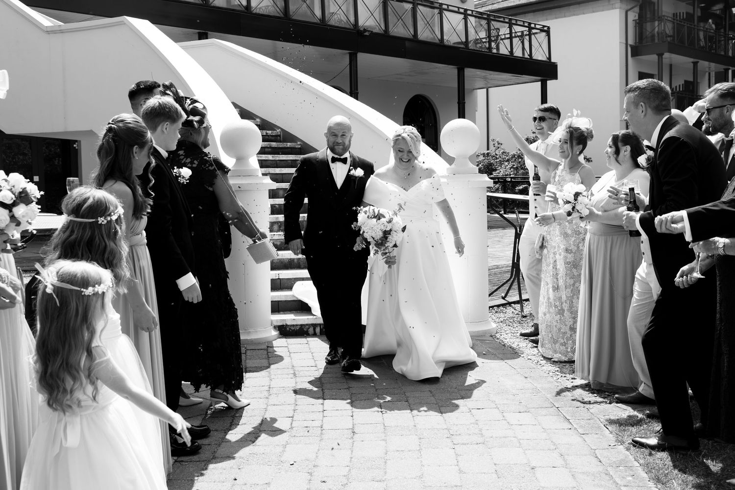 Black and white photo of a bride and groom walking through a confetti-throwing wedding ceremony exit.
