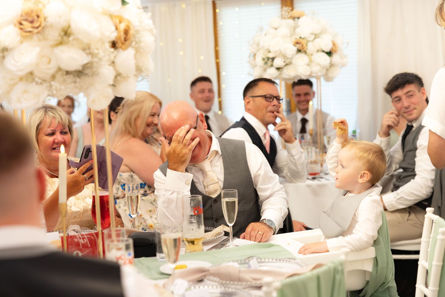 Wedding party and guests share laughter during speeches at a beautifully decorated reception.