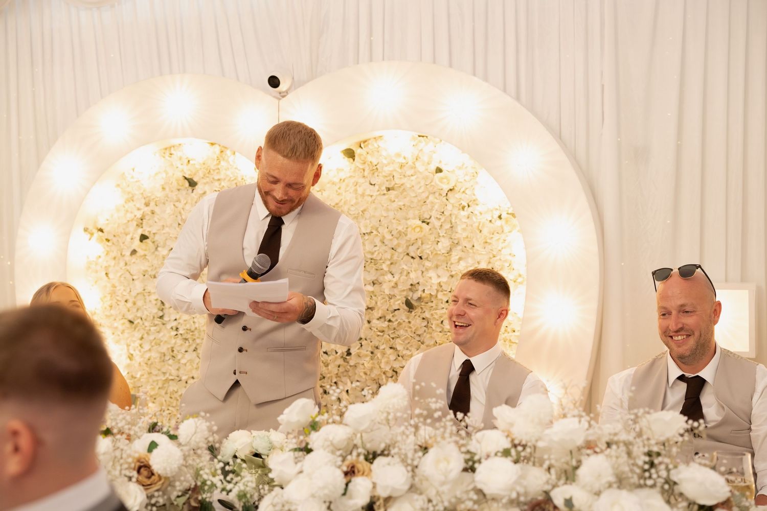 Groom reading speech at wedding reception with floral circular backdrop and laughing groomsmen.