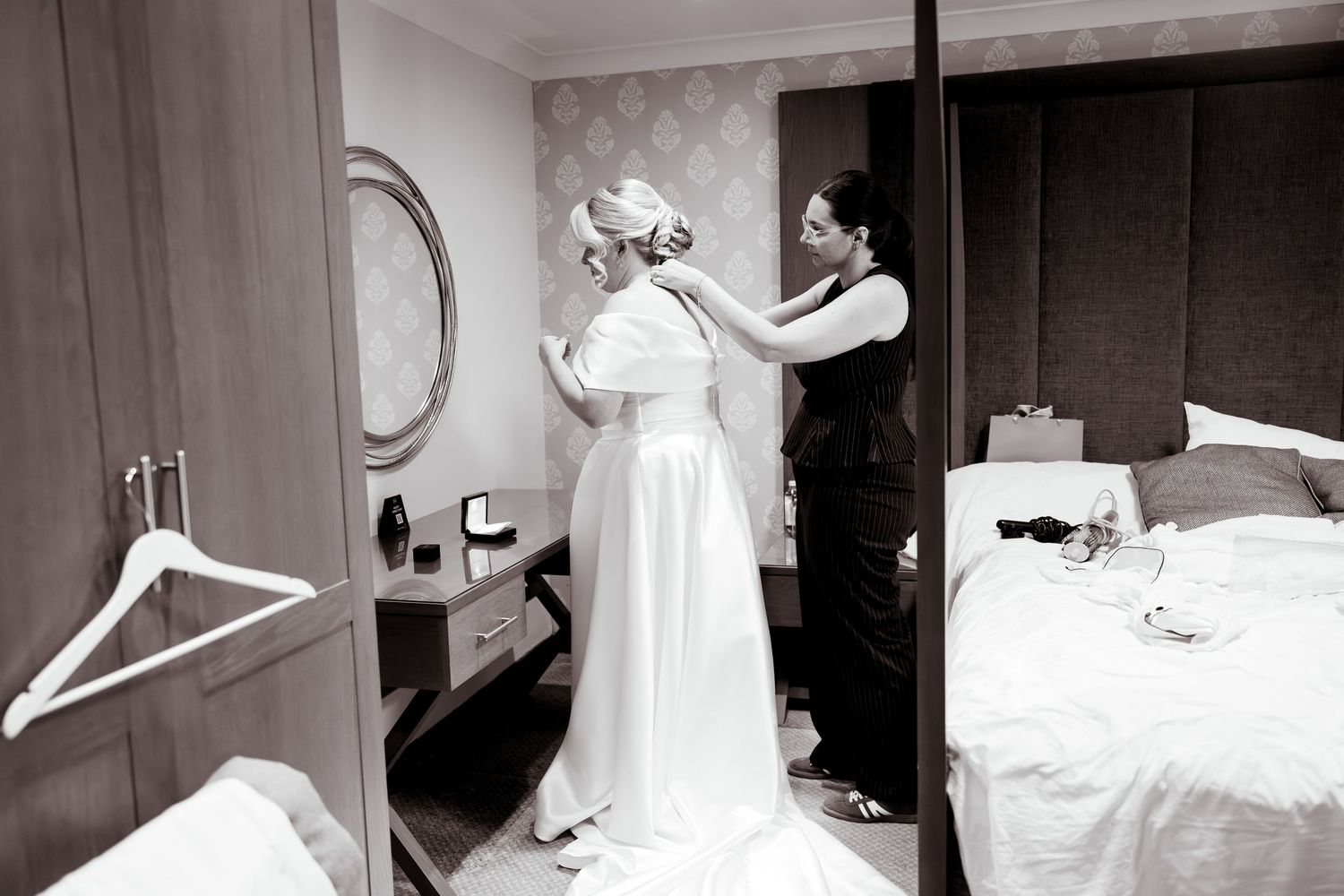 Black and white photo of a bridesmaid helping a bride into her wedding dress in a hotel room.