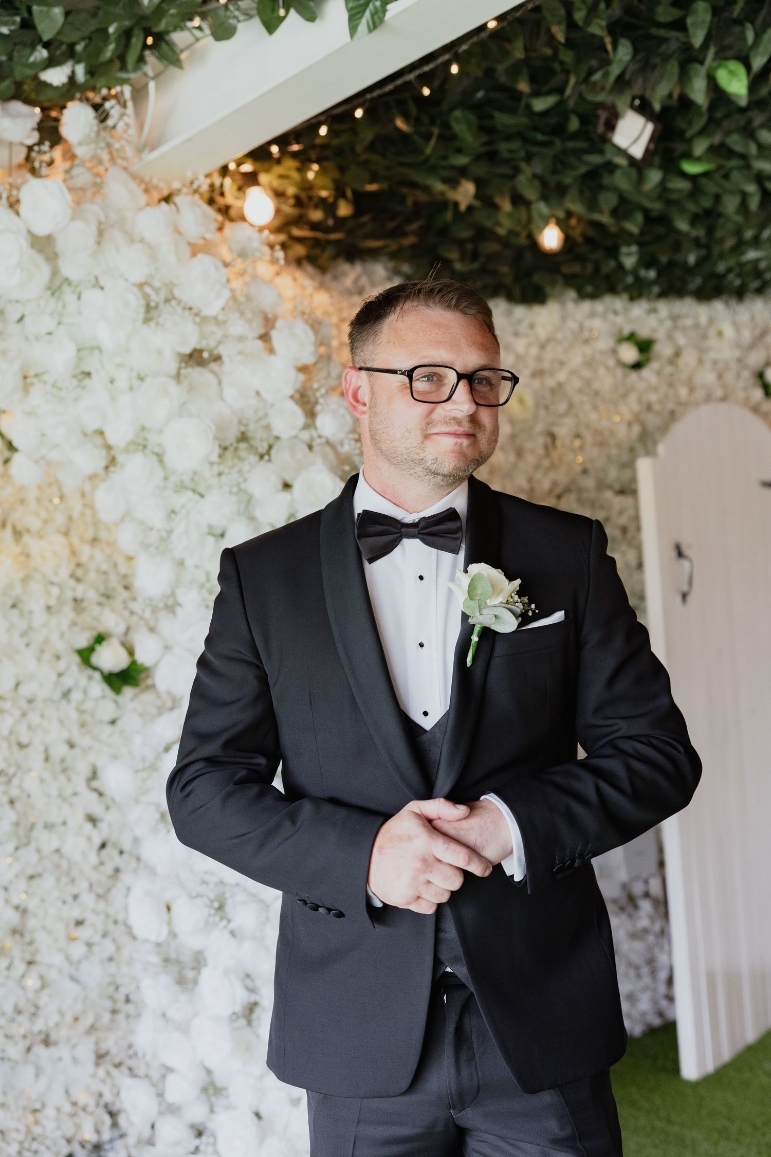 A dapper groom in a black tuxedo with bow tie and boutonniere poses before a white floral wall.