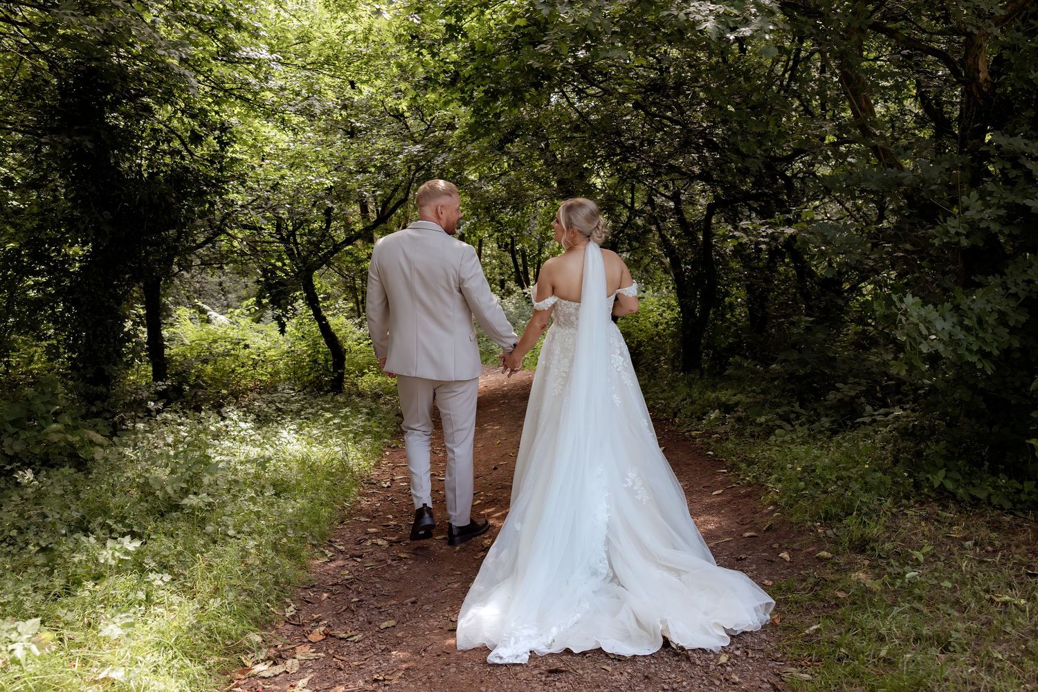 Bride and groom walking hand in hand along a lush green woodland path on their wedding day.