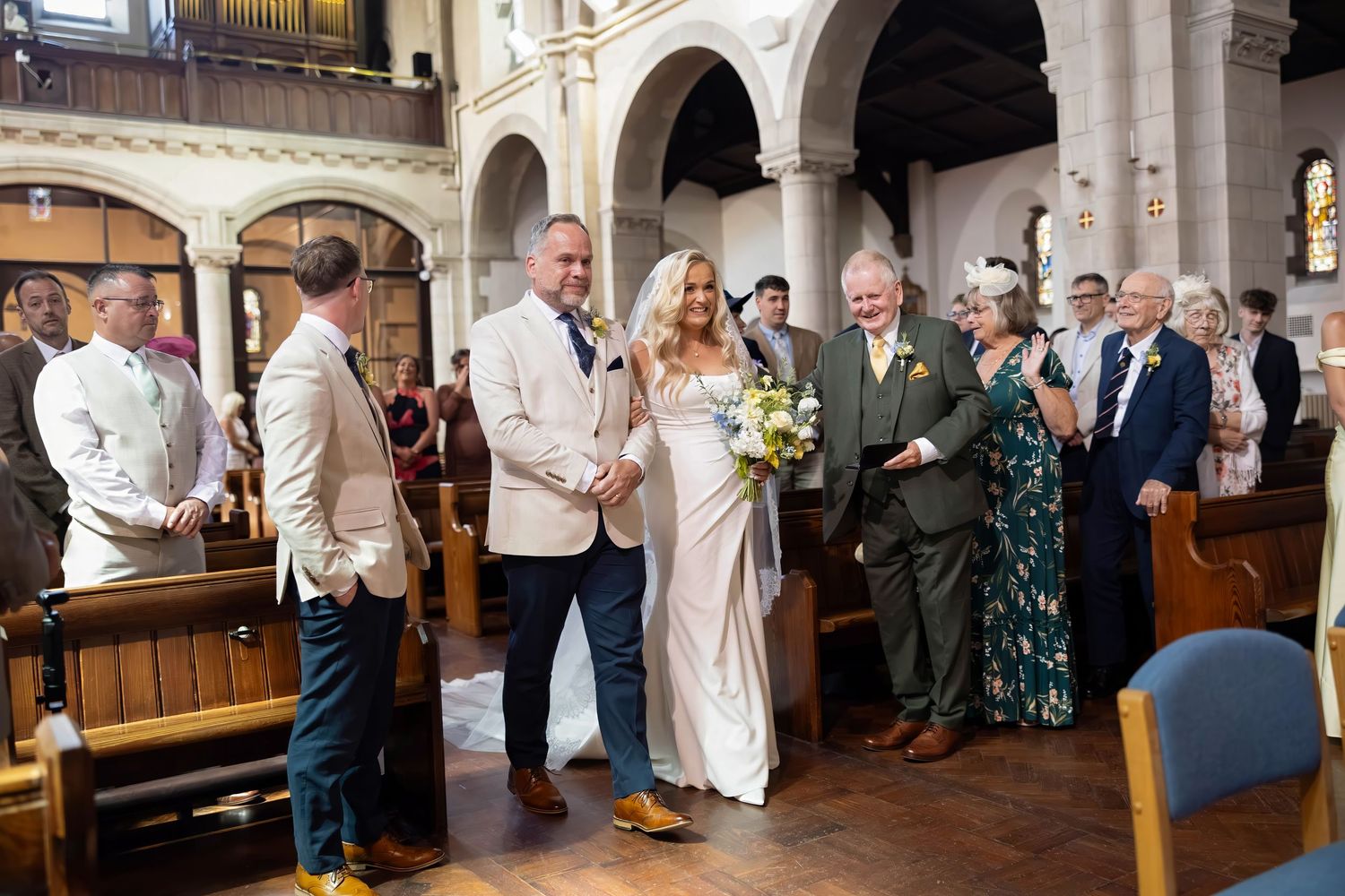 Radiant bride walking down church aisle with groom and father during traditional wedding ceremony.