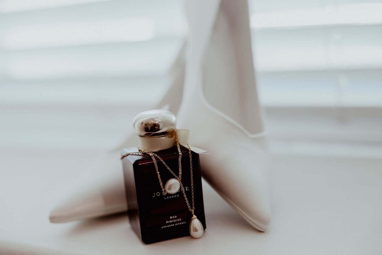 Elegant bridal accessories featuring white heels, gold necklace with pearl pendant, and dark perfume bottle on white surface.