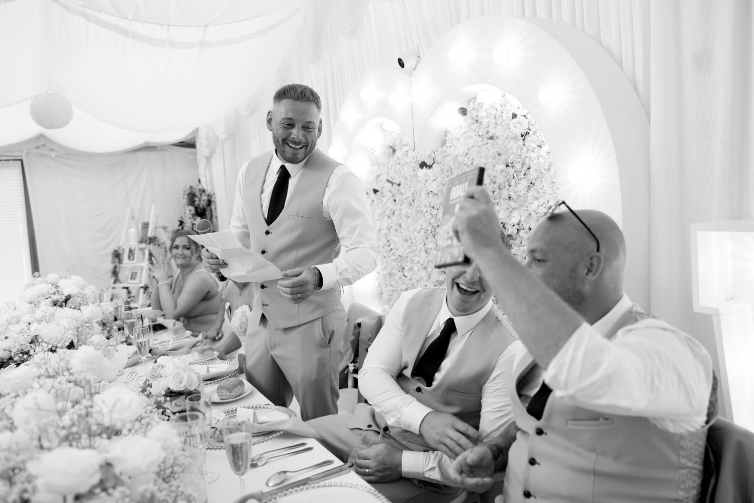 Black and white photo of groom giving wedding speech to laughing guests at head table.