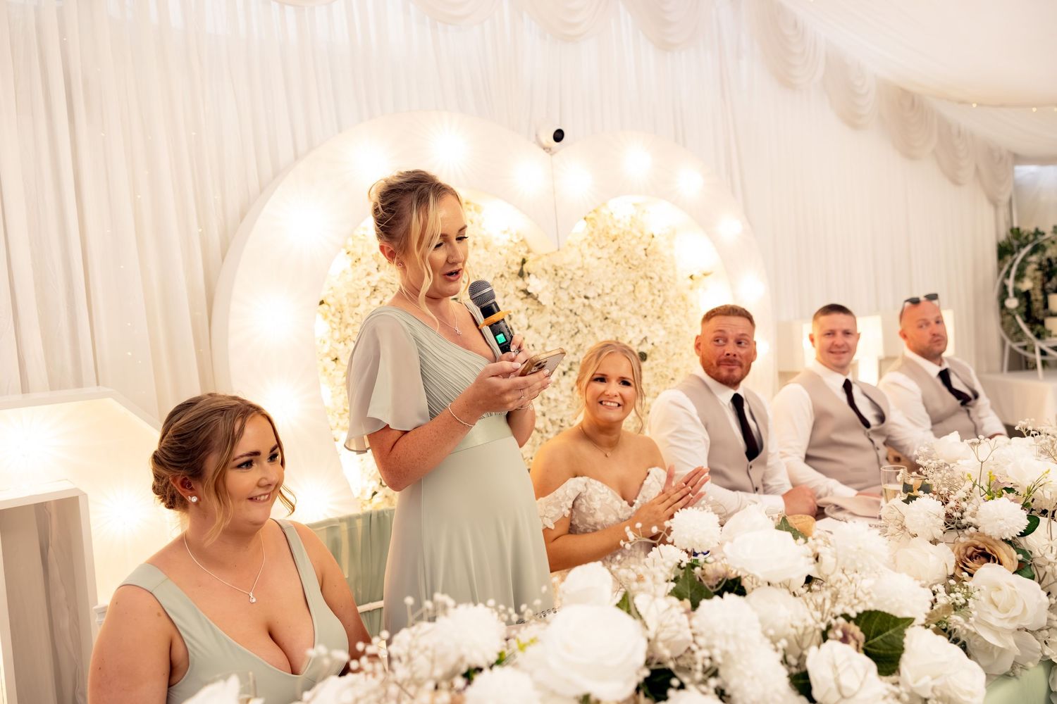Bridesmaid delivers a touching speech as wedding party smiles at a beautifully decorated reception.