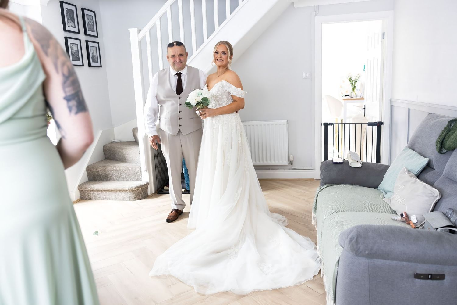 Bride in flowing white gown with train stands with escort in grey vest at home.