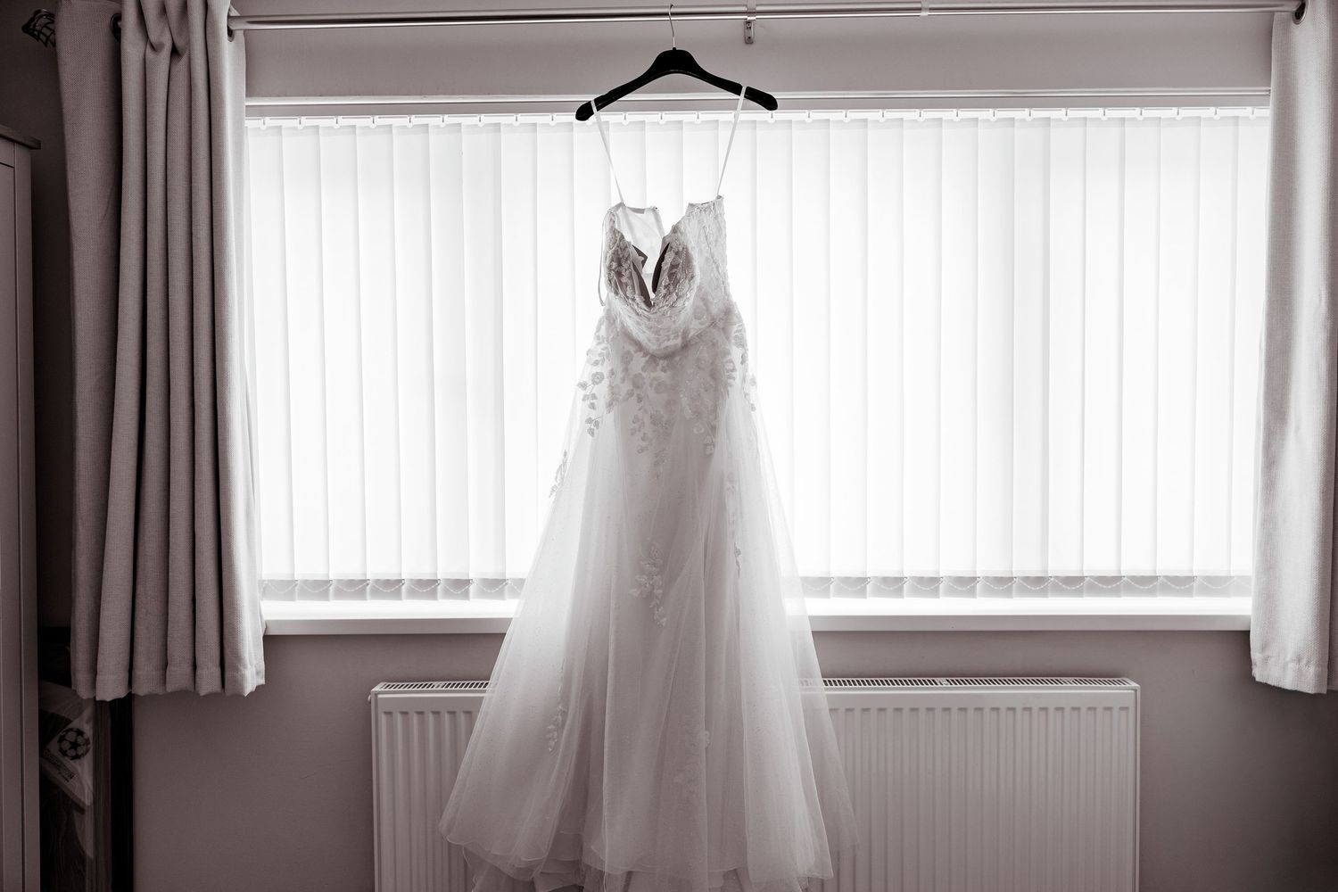 Elegant lace and tulle wedding dress hanging in front of a window with white vertical blinds.