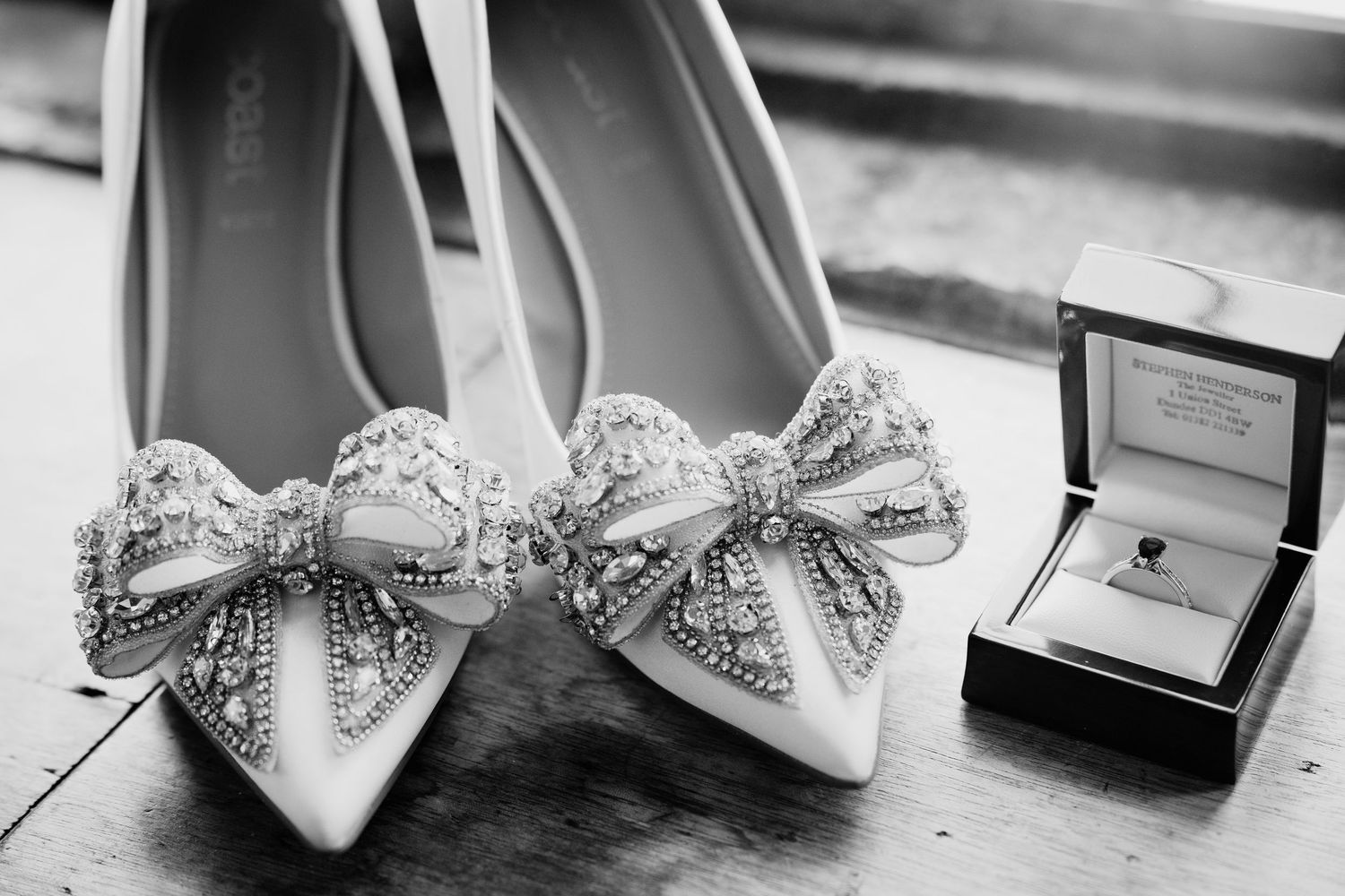 Black and white photo of jeweled bow bridal heels beside a diamond engagement ring in a box on a wooden surface.
