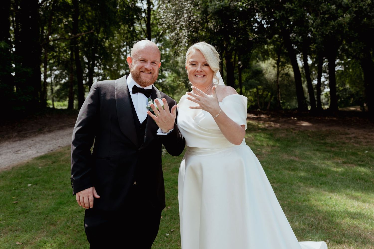 A newlywed couple in wedding attire smiling and showing off their wedding rings outdoors.