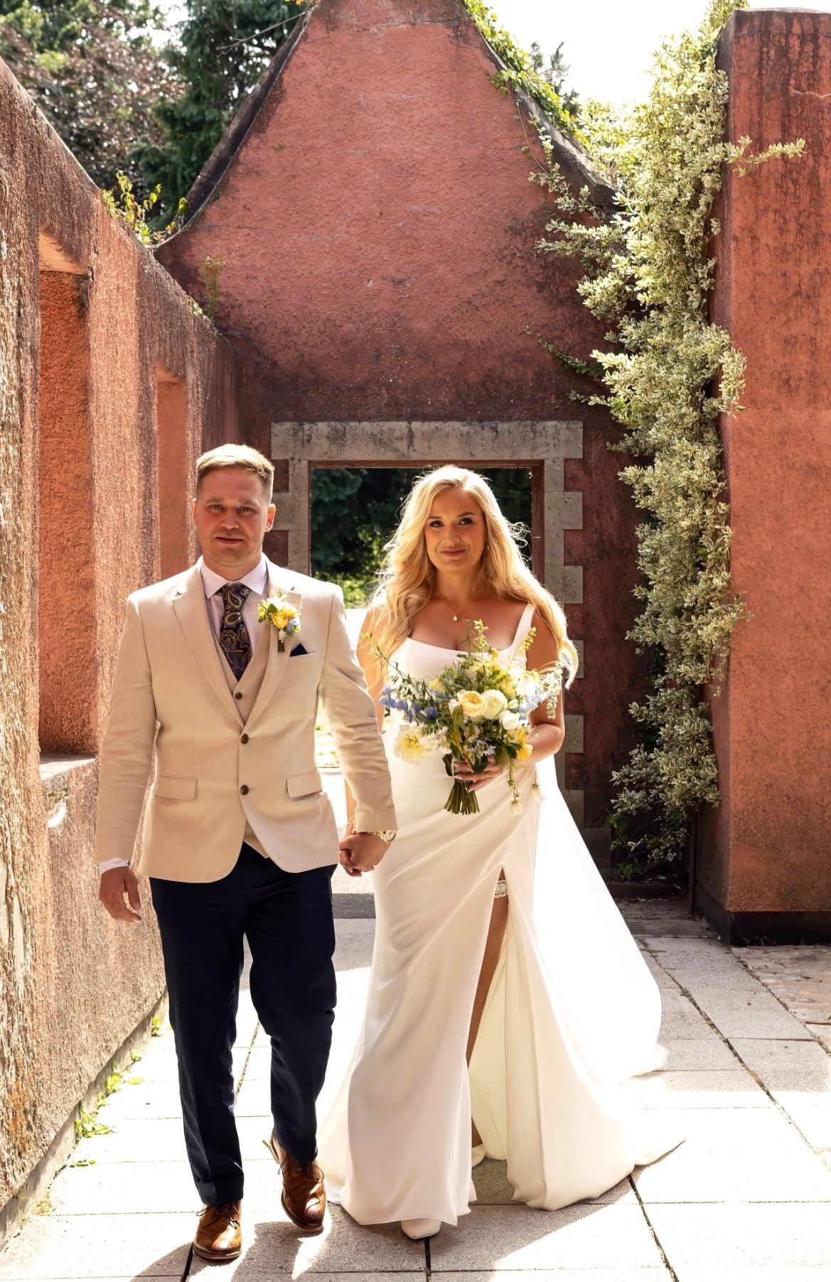 Bride and groom walk hand-in-hand through rustic red stone archway, bride holding yellow and blue bouquet.