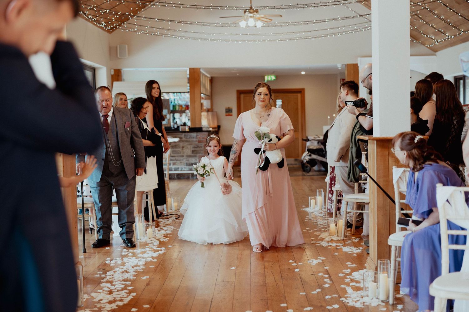 Bridesmaid in pink dress walks flower girl down petal-lined wedding ceremony aisle.