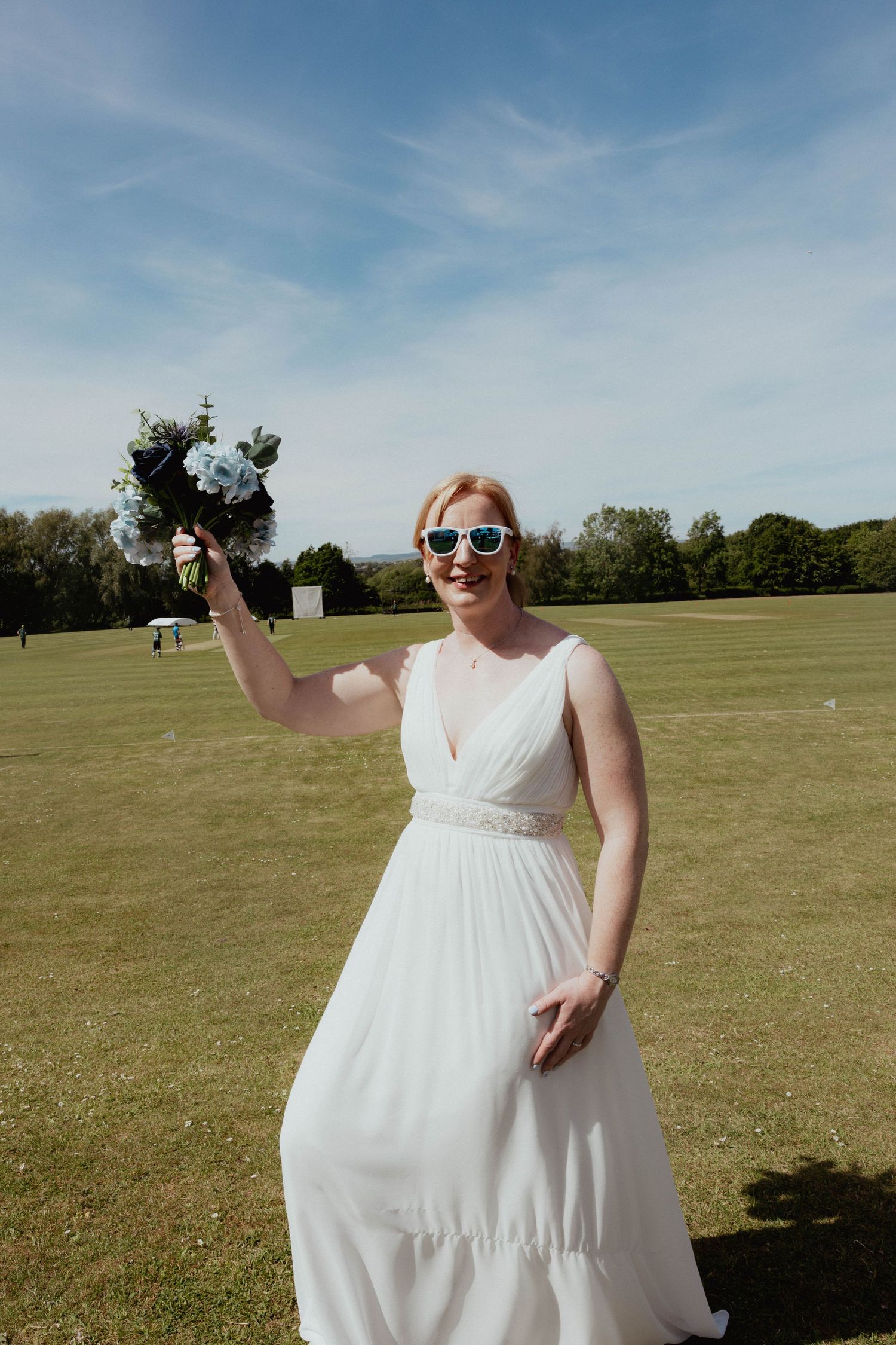 Radiant bride in a white gown and sunglasses raises her blue bouquet outdoors on a sunny day.