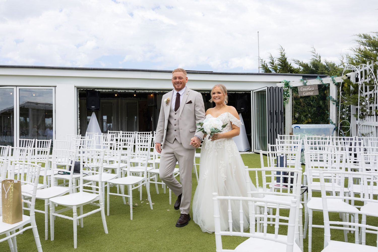 Bride and groom walking together outdoors at a wedding venue with white chairs arranged on green grass.