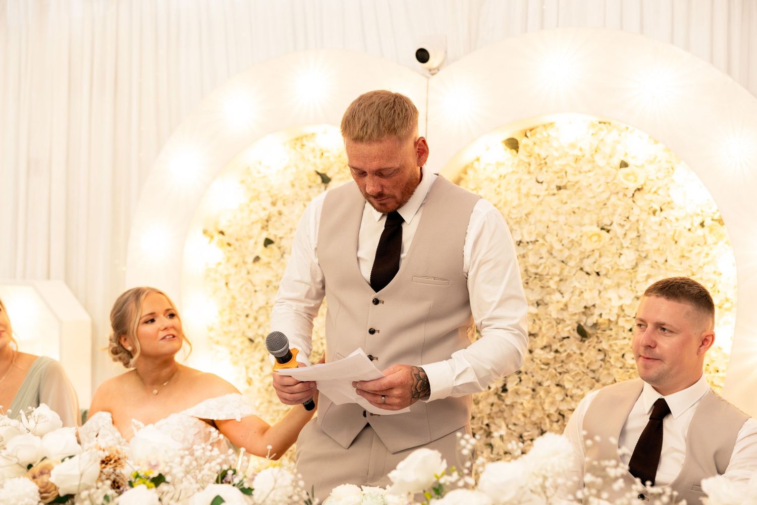 Best man delivers a heartfelt wedding speech at an elegant white floral reception venue.
