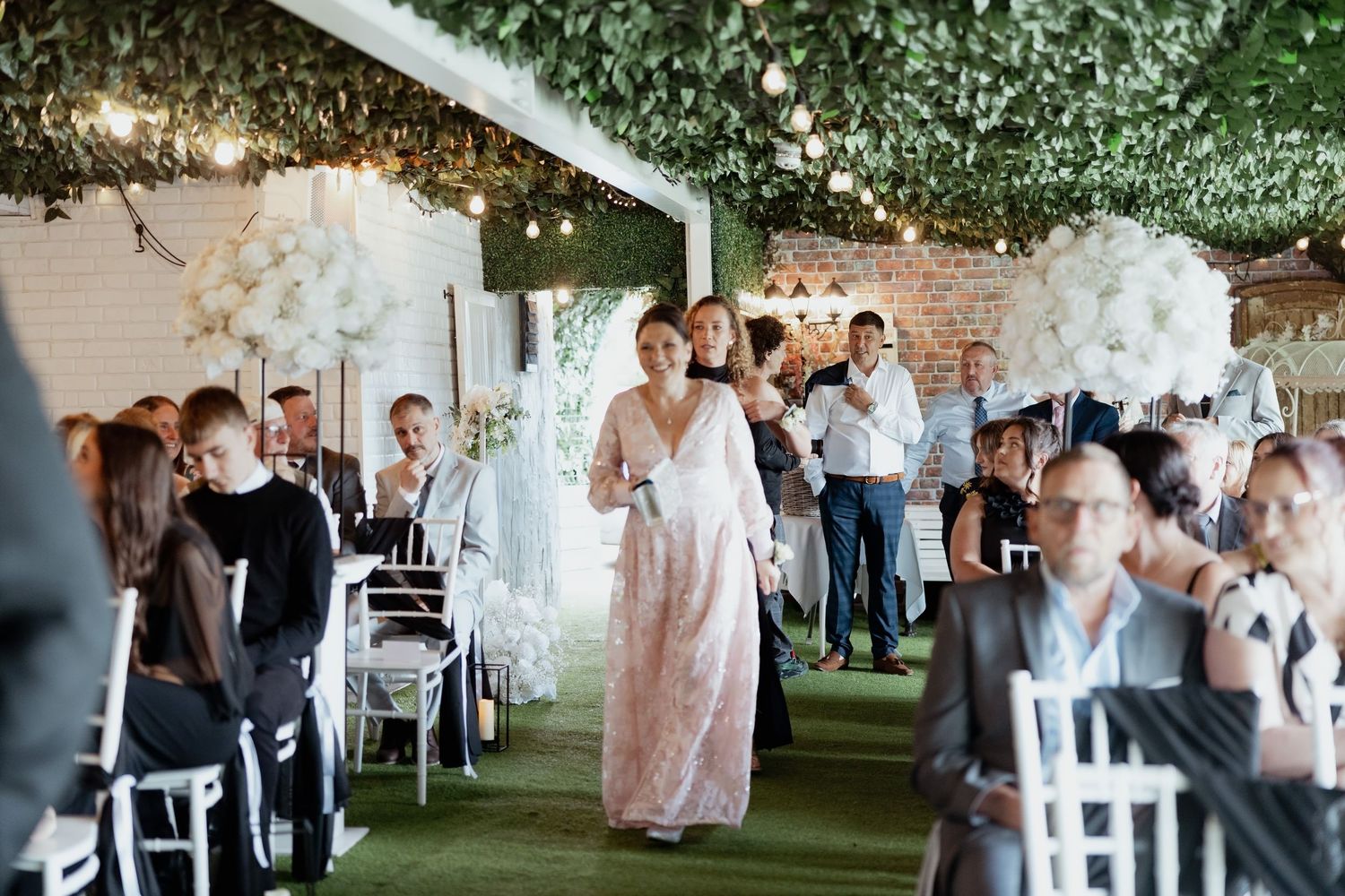A woman in a pink lace dress walks down the aisle at an elegant green and white decorated wedding venue.