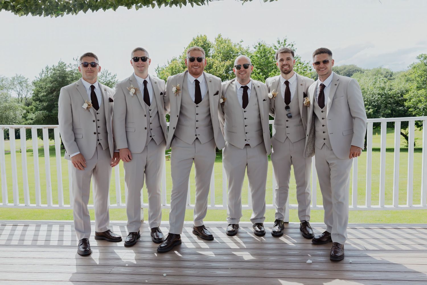 Six groomsmen in matching light grey suits with dark ties and boutonnieres posing on a wooden deck at a wedding.