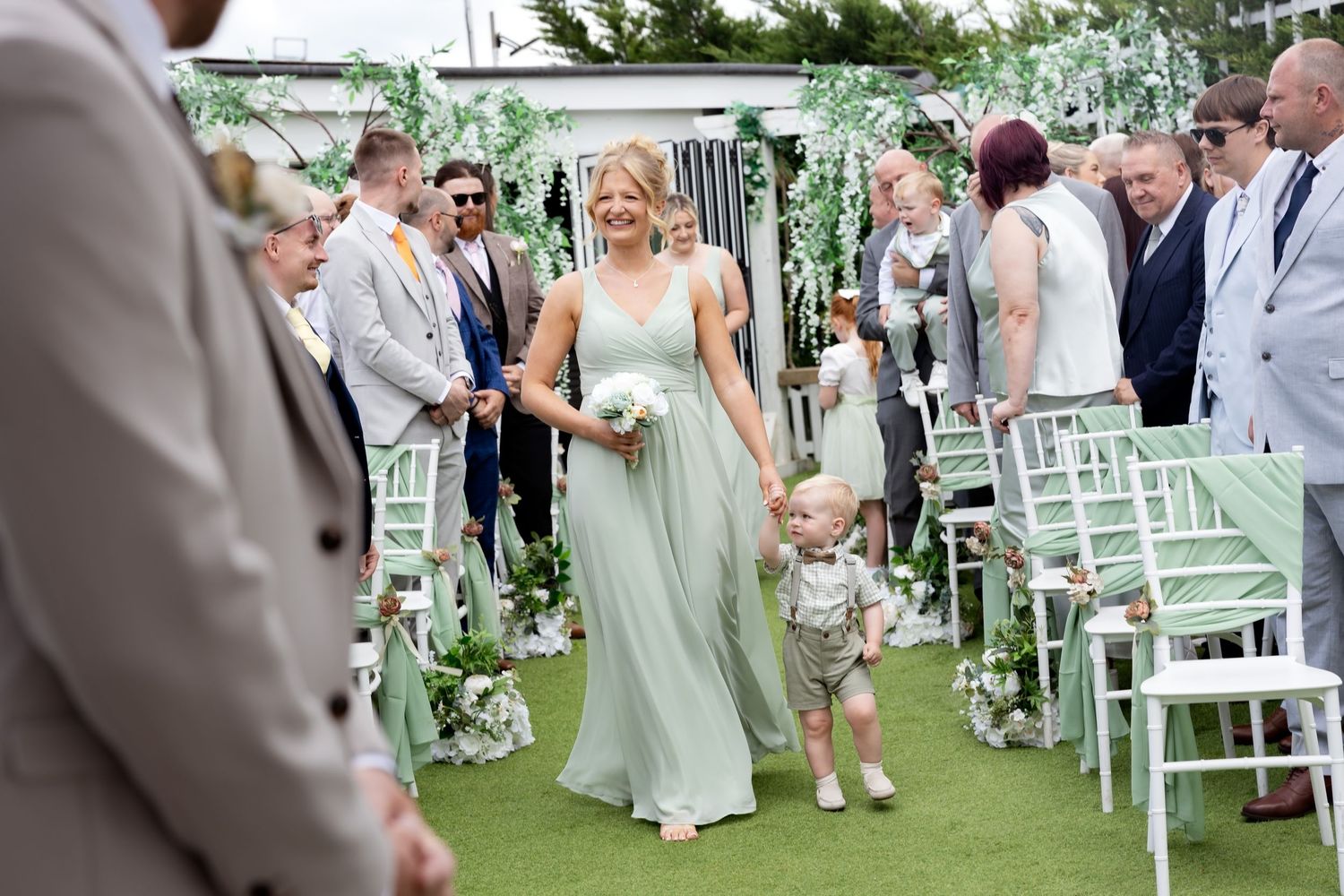 Bridesmaid in sage green dress walks down aisle with young child at outdoor wedding.