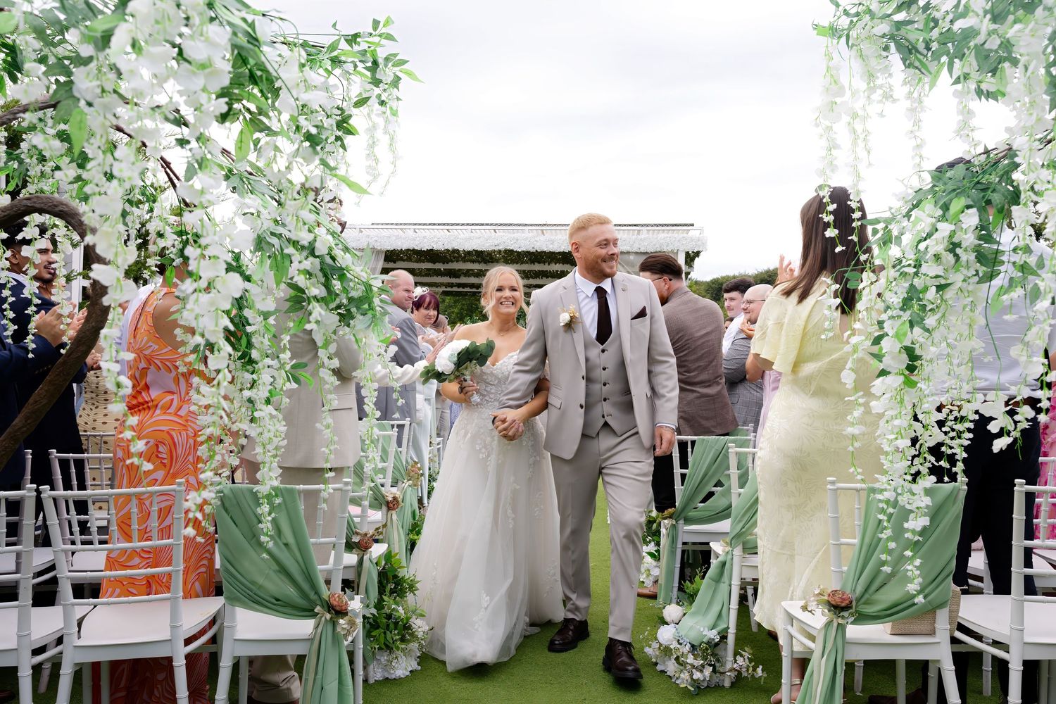 Bride and groom walk down outdoor wedding aisle lined with white floral arches and green chairs.