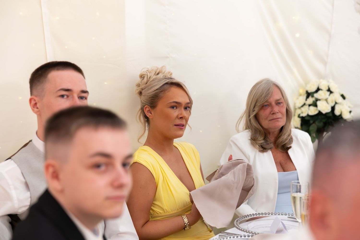 Wedding guests in yellow dress and white jacket listening attentively during reception speeches.