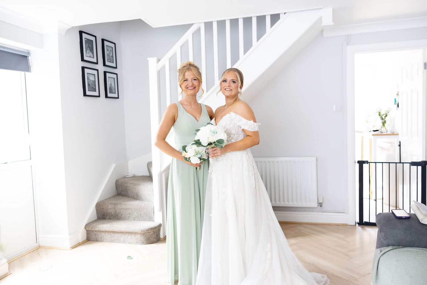 Bride and bridesmaid in sage green dress smiling together near white staircase.