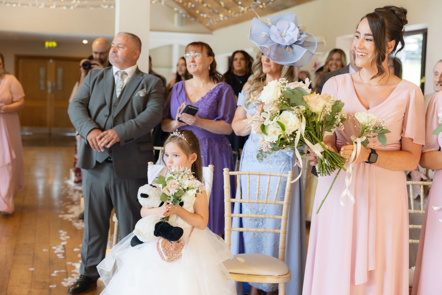 Flower girl and bridesmaid holding bouquets stand during wedding ceremony in elegant venue.