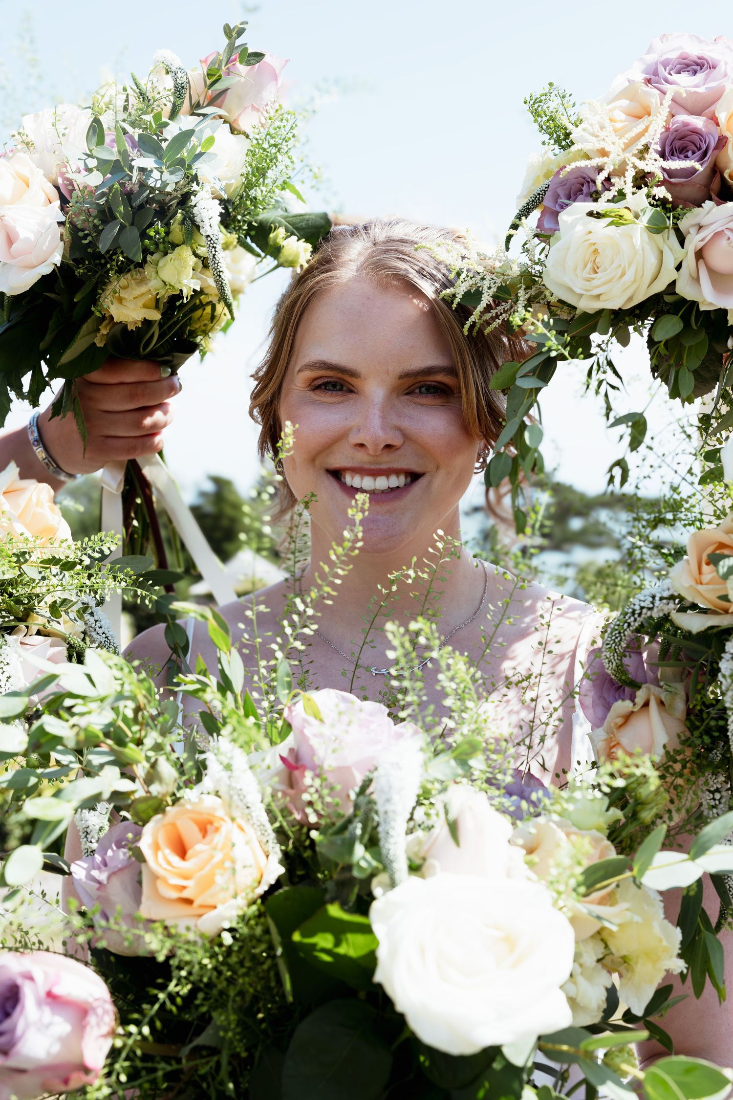 Smiling bridesmaid surrounded by lush pastel wedding bouquets of roses and greenery on a sunny day.