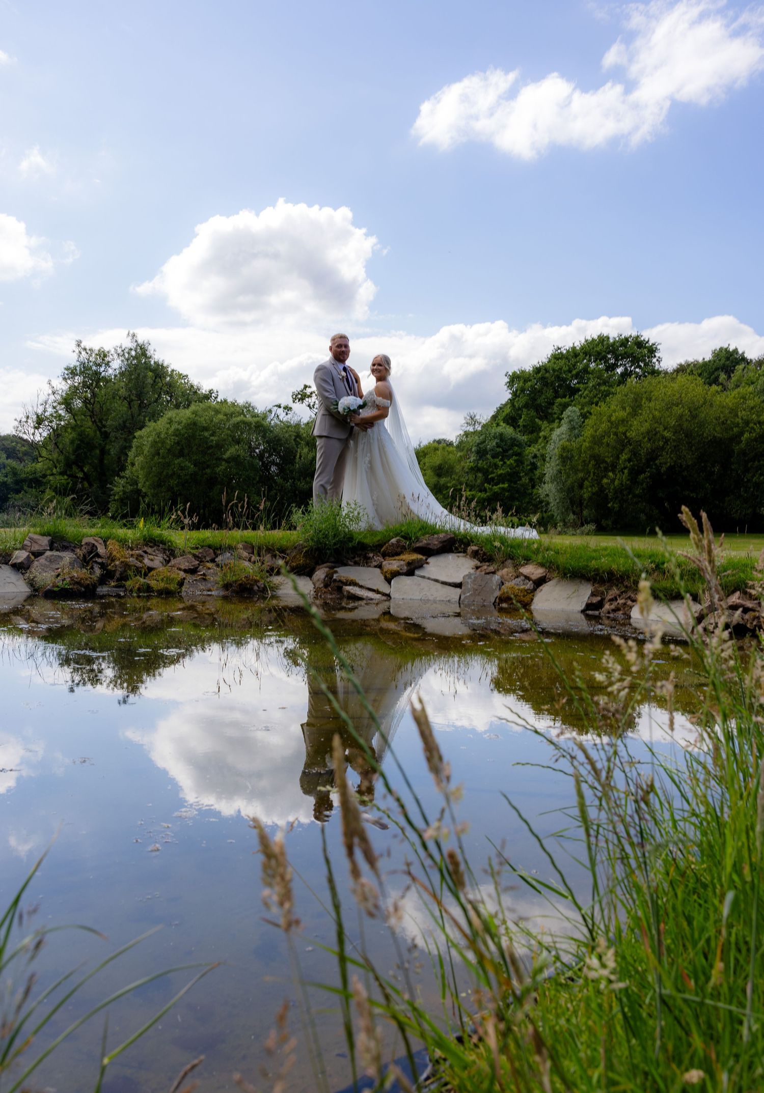 Bride and groom standing by a reflective pond surrounded by lush greenery on their wedding day.