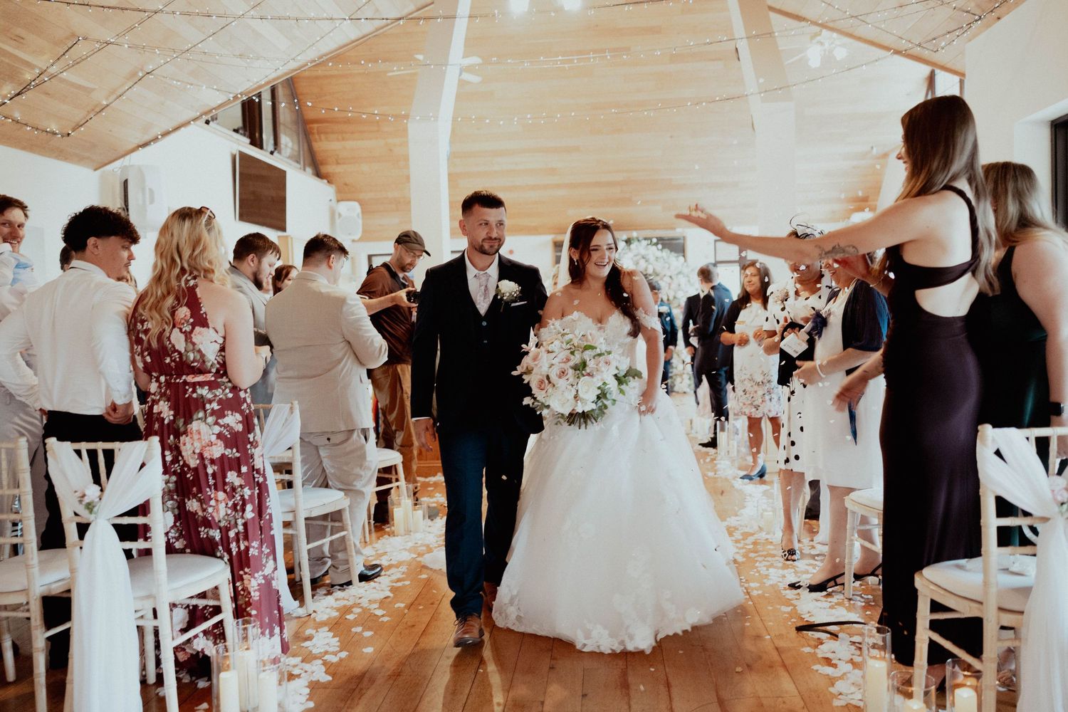 Newlyweds walk down the aisle as guests celebrate with confetti in a beautiful rustic barn venue.