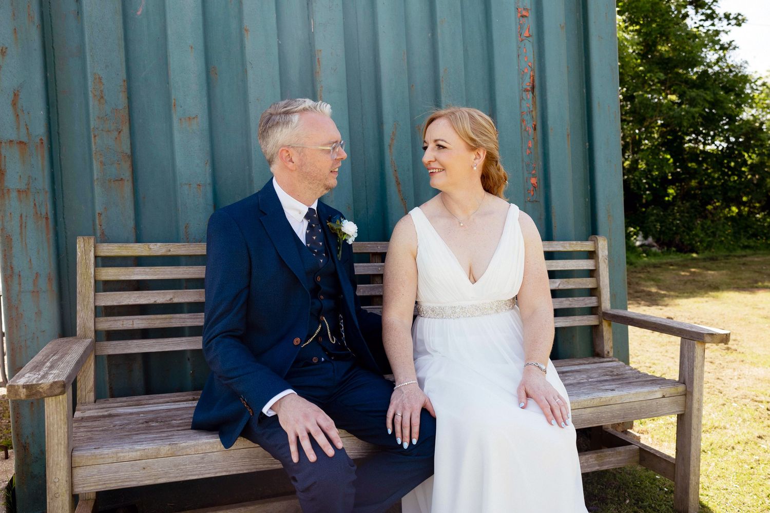 Bride and groom sit together on a wooden bench smiling at each other on their sunny wedding day.