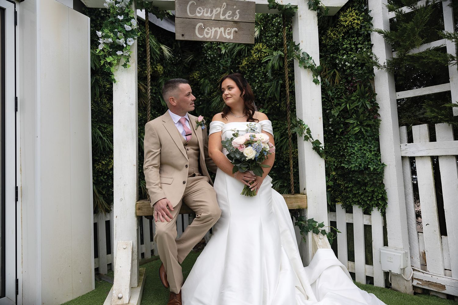 Bride and groom pose under a Couples Corner sign at a wedding venue with white picket fence and green foliage backdrop. welsh weddings