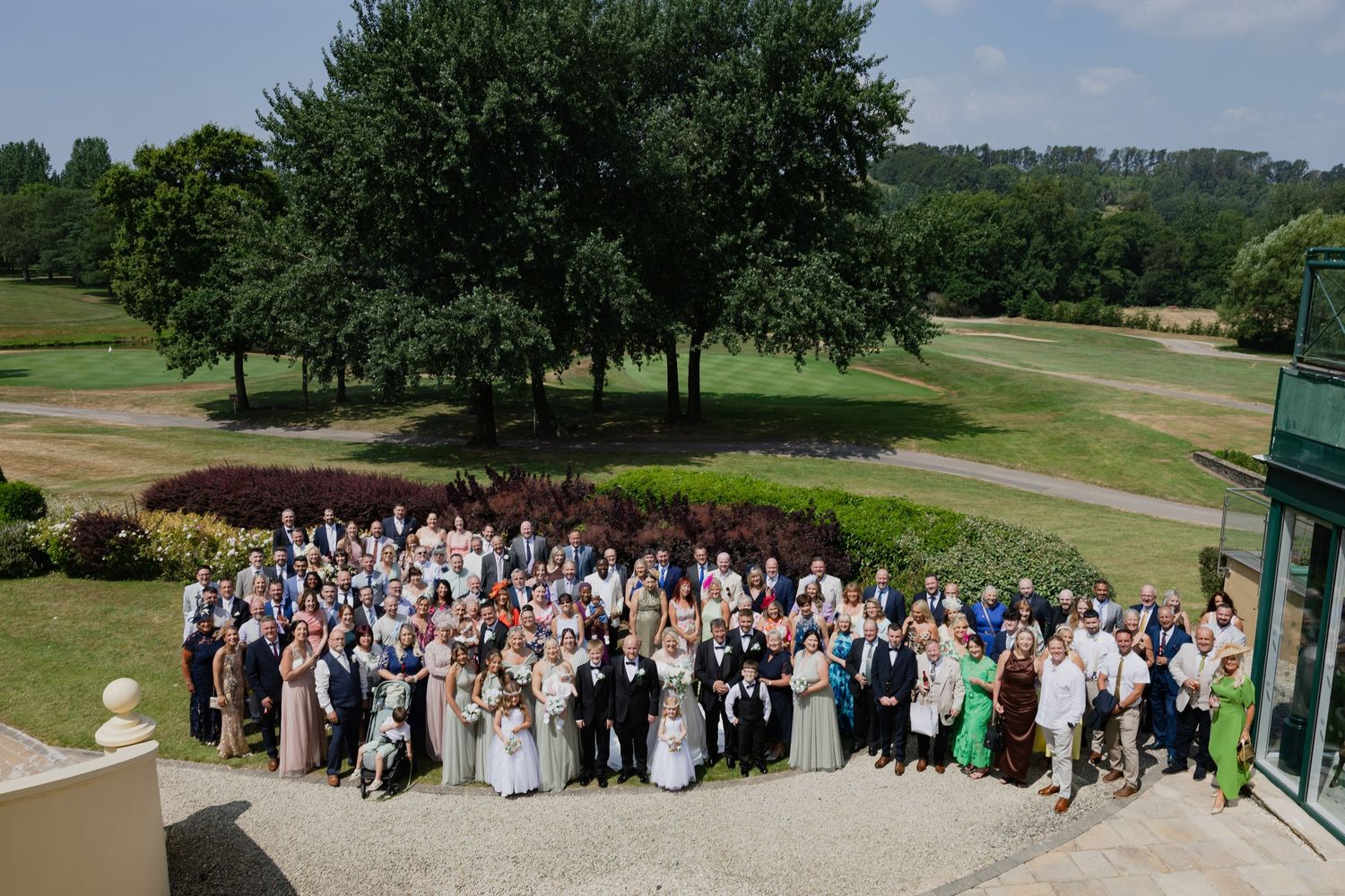 Large wedding group photo outdoors at a golf club venue, with guests gathered on a terrace surrounded by lush trees.