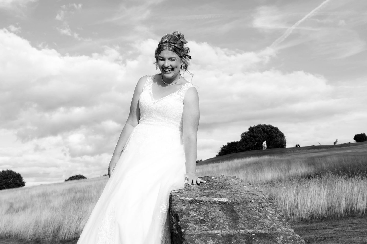 Bride in white gown smiling outdoors in black and white photo with cloudy sky and field backdrop.