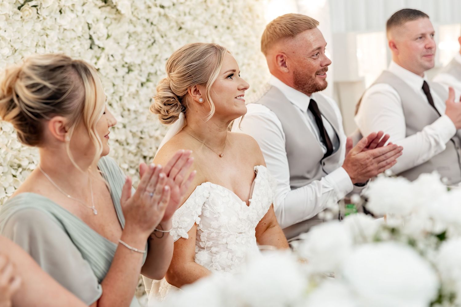 Bride in lace gown and wedding party applauding at elegant white floral decorated wedding reception.